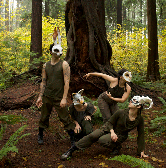 Four dance artists pose in the forest. They are wearing all green and white animal masks.