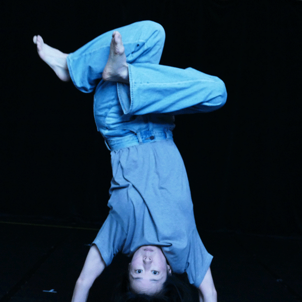 A dance artist wearing all blue stands on her hands with her legs crossed over each other.