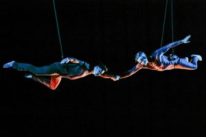 Two aerial dance artists hang horizontally with their left hands clasped.