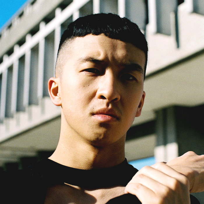 A dance artist stands outside in front of a concrete building. Here is wearing a dark shirt and is holding his hands up near his neck.