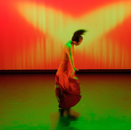 A dance artist mid performance. There is a orange and yellow background behind her and the floor is lit with green light. The dancers hair is blurry with movement and her arms are extended as if she is about to raise them up.