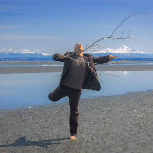 A dance artist stands on a beach in front of water and snowy mountains on a sunny day. He is wearing a dark grey blazer and pants with a light grey shirt under. He has one leg out and his head back and is holding a long multi branched stick.