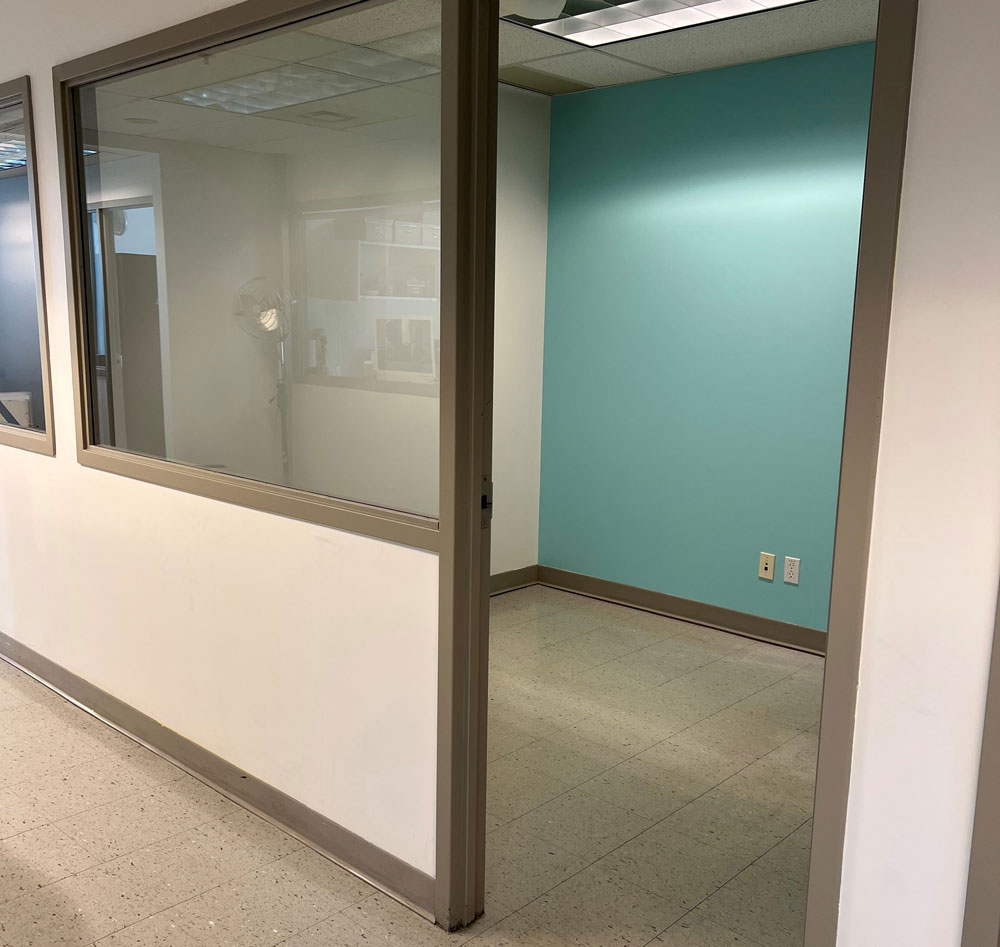 An empty office space viewed through an open door, with a window and a turquoise rear wall.